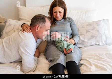 A couple sits on the bed, adoring their peaceful sleeping newborn cradled in the mother's lap, sharing a quiet family moment Stock Photo