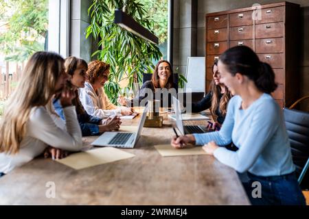 A group of smiling women engaged in a meeting at a wooden table in a modern office setting, illustrating teamwork and female entrepreneurship. Stock Photo
