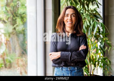 Confident woman entrepreneur with arms crossed stands in an office environment, smiling and looking at the camera. Stock Photo