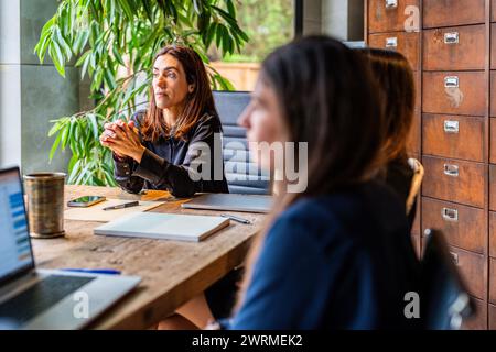 Two female colleagues engage in a focused conversation, one woman looking at the other who is facing away from the camera. Stock Photo