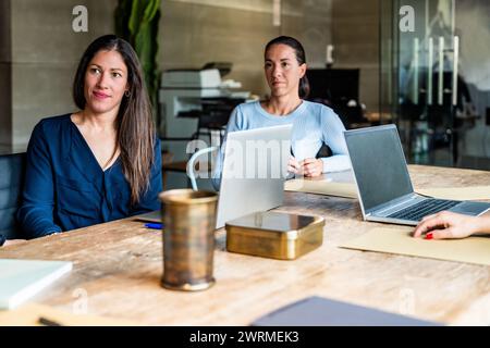 Two women collaborating at a wooden table in a modern office, one looking at the camera. Stock Photo