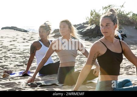 Three women practicing yoga on the beach at sunset, engaged in a seated twist pose. Stock Photo