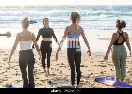 A group of women participates in a peaceful yoga class on the beach in the background, standing in Tadasana, or Mountain Pose. Stock Photo