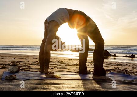 Practicing an advanced yoga backbend, the Wheel Pose, during a serene sunset on the beach. Stock Photo