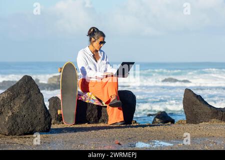 A focused digital female nomad woman sits on a rocky shore, using her laptop with a skateboard beside her against a backdrop of a lively ocean in a Fu Stock Photo