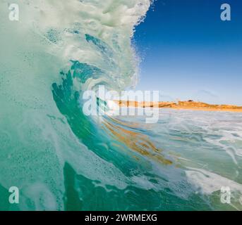 A stunning view from inside a curling turquoise wave, capturing the dynamic textures as it breaks towards a sunlit beach Stock Photo