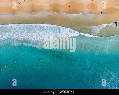 A top-down shot of turquoise waves rolling onto a golden sandy beach with swimmers and sunbathers enjoying the coastal beauty Stock Photo
