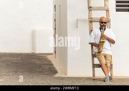 A bald male sits on a wooden ladder outside, playing a golden saxophone, contributing to the celebration of International Jazz Day. Stock Photo