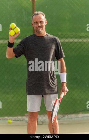 An active Mature man stands on a tennis court, holding tennis balls and a racket, looking at the camera with a smile. Stock Photo