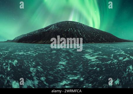 A spectacular display of Aurora Borealis illuminates the sky above a snowy, icy terrain in Iceland, with a mountain as backdrop. Stock Photo