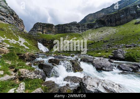 Swift mountain stream flows through a rocky landscape, with a waterfall backdrop in the verdant Valle de Ordesa, Huesca Stock Photo