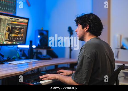 A focused music producer edits audio tracks on a digital workstation with a MIDI keyboard in a home studio setting. Stock Photo