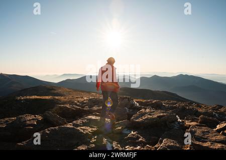 Back view of an unrecognizable lone hiker stands facing the rising sun on a rocky mountain peak, overlooking layered hills stretching into the distanc Stock Photo