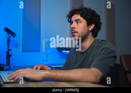 A focused man engaged in a sound editing session in a studio, illuminated by blue ambient light, with a microphone and laptop in front of him. Stock Photo