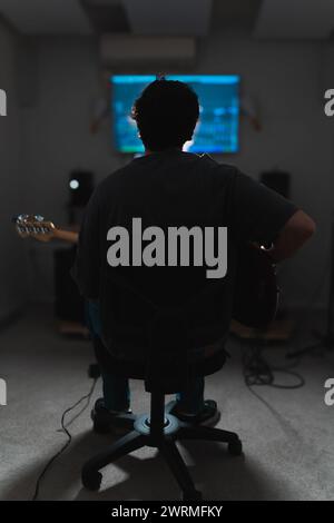 Back view of unrecognizable male musician sits with a guitar in a dimly lit sound studio, facing audio mixing equipment during a recording session. Stock Photo