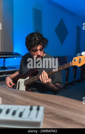 A focused male musician expertly plays a bass guitar during a sound studio recording session, with music equipment in the background. Stock Photo