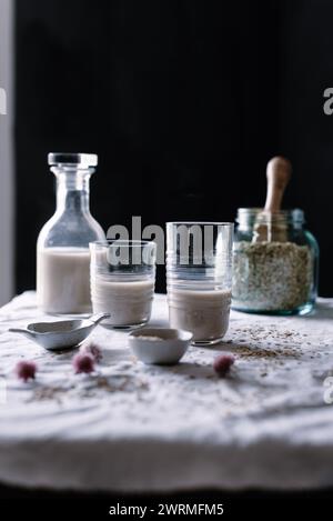 A cozy, rustic scene featuring homemade oat milk in clear glasses, with a bottle and jar of oats on a linen-covered table Stock Photo