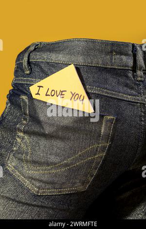 A heartfelt love note on a yellow sticky paper is subtly tucked into the back pocket of a denim jean against a yellow background Stock Photo