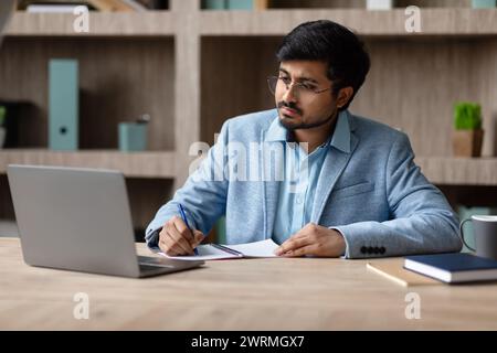 Hindu businessman sits at laptop computer taking notes at office Stock Photo