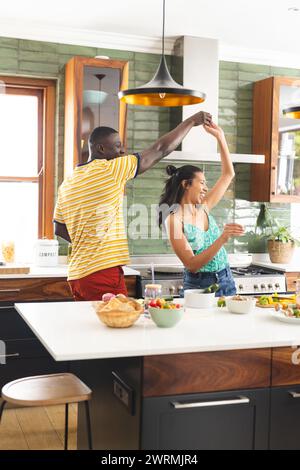Young African American man and biracial woman dance joyfully in a sunlit kitchen Stock Photo