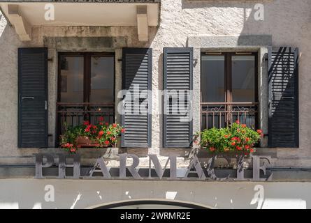 Facade of an old building with the metal sign of a pharmacy under a pair of windows with blooming geraniums, Chamonix, Haute Savoie, France Stock Photo