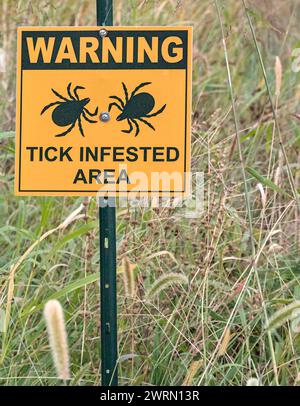 warning tick infested area sign in a grassy field, public park (lyme disease prevention) photo, tall grass, deer, dog, lone star ticks ground Stock Photo