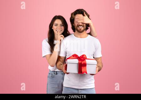 Surprised bearded man in a white shirt unknowingly holds a gift with a red ribbon as a smiling woman Stock Photo