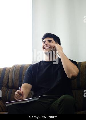 Image of a young man sitting on his couch, smiling as he works from home. He is seen writing in a notebook while holding a call on his cell phone Stock Photo