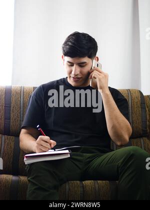 Young Caucasian man talks on cell phone while taking notes, sitting in a home office or living room Stock Photo