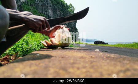 View of an African man with a machete cutting a coconut, with Santa Catarina tunnel as a background in Sao Tome, Africa Stock Photo