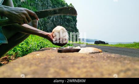 View of an African man with a machete cutting a coconut, with Santa Catarina tunnel as a background in Sao Tome, Africa Stock Photo