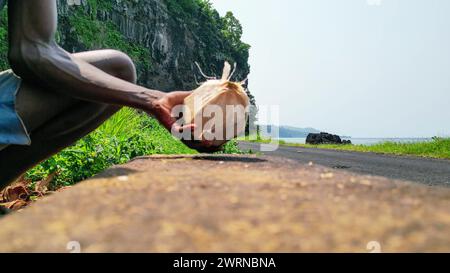 View of an African man with a machete cutting a coconut, with Santa Catarina tunnel as a background in Sao Tome, Africa Stock Photo