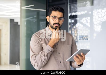 Pensive businessman holding a digital tablet, deep in thought inside a contemporary corporate environment. Stock Photo