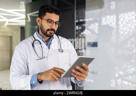 Focused male doctor in lab coat and stethoscope using a digital tablet in a modern hospital setting. Stock Photo