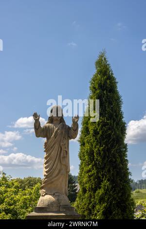 Statue of Jesus Christ as a headstone on a grave. Typical gesture. Cemetery beside All Saints church. High bright green conifer growing beside. Blue s Stock Photo