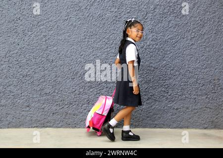 4 year old Latina brunette girl with eyeglasses, uniform and backpack walks to school this back to school Stock Photo