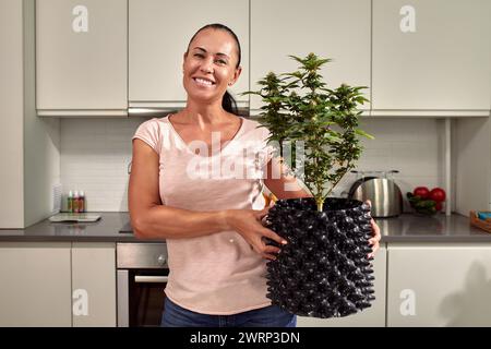 An image showing an adult female in casual wear, holding a potted plant in a modern kitchen setting. The focus is on the homely atmosphere and indoor Stock Photo