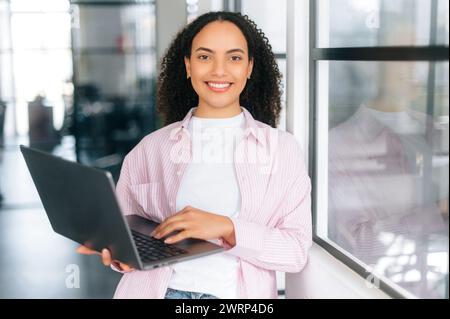 Successful brazilian or hispanic stylish curly haired young woman, software engineer, digital manager, stands in a modern office with an open laptop in hands, looking at the camera, smiles friendly Stock Photo