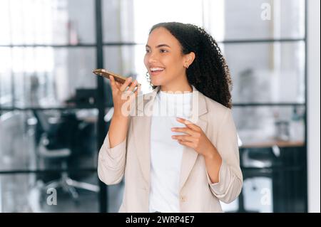 Excited happy brazilian or latino elegant curly haired woman, secretary, company ceo, stand in a modern office space, using mobile phone, recording audio message, speaks on speakerphone, smiles Stock Photo