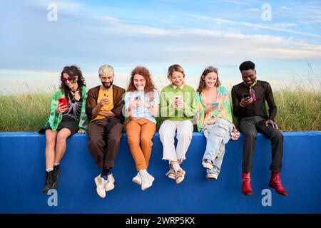 Happy people in nature sitting on blue wall, looking at phones under blue sky Stock Photo