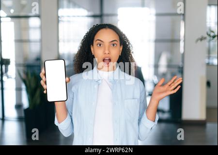 Surprised brazilian or hispanic curly haired woman in a pastel blue shirt, stands in a modern office, showing smart phone with white empty mock-up screen, shocked looks at camera. Copy-space concept Stock Photo
