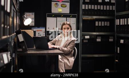 Confident female private detective investigator reviews clues in office, searching on laptop for evidence to solve crime. Profile portrait of focused caucasian policewoman using personal computer. Stock Photo