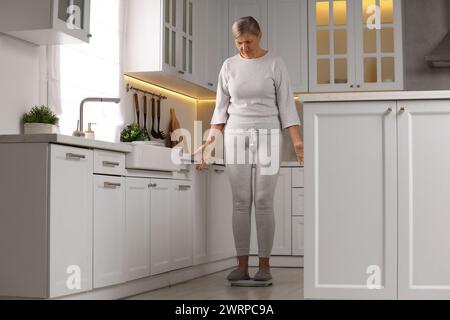Menopause, weight gain. Concerned woman standing on floor scales in kitchen Stock Photo