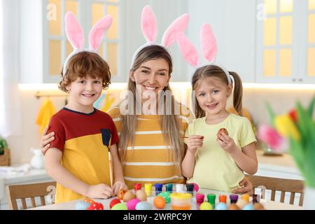Painting Easter eggs. Happy mother and her cute children with bunny ears at table in kitchen Stock Photo
