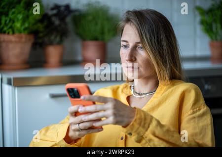 Serious woman holding smartphone attentive reading received message sitting at home with houseplants Stock Photo