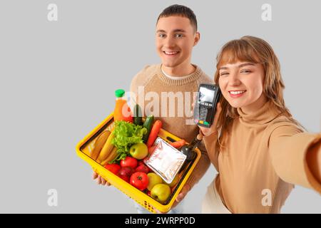 Young couple with full shopping basket and payment terminal taking selfie on light background Stock Photo