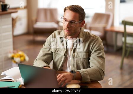 Portrait of young concentrated man in glasses, working from coffee shop, studying, looking thoughtful while completing task on laptop, making notes in Stock Photo