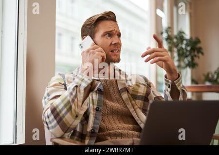 Image of man feeling stressed while talking over the phone. Businessman sitting in cafe with laptop, answering a call, hear bad news over the Stock Photo