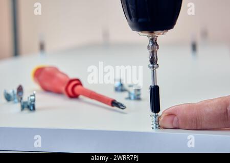Furniture connector fitting, connecting dowel for cam lock nut, assembler screws connection screw into chipboard panel using an electric screwdriver. Stock Photo