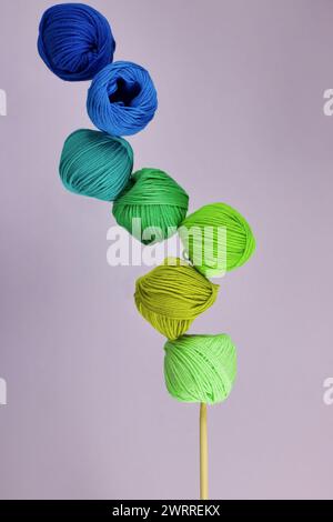 group of cotton balls yarn in the shades of green and blue in balance on wooden knitting needle, lilac background Stock Photo